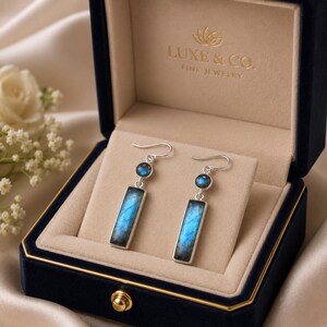 Puede incluir: Un par de pendientes colgantes de plata con una pequeña piedra de labradorita redonda sobre un colgante rectangular de labradorita. Los pendientes se presentan en un joyero de terciopelo azul marino con el texto LUXE & CO. FINE JEWELRY.