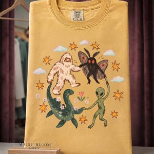 May include: A mustard yellow t-shirt featuring a graphic print of a white Bigfoot, a black mothman, a green sea monster, and a green alien holding hands in a circle. The design includes orange stars, light blue clouds, and small pink flowers.