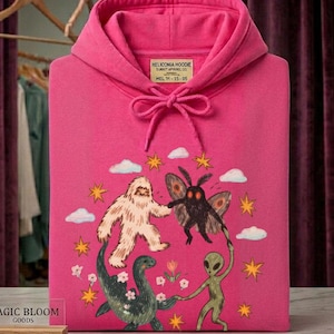 May include: A bright pink hooded sweatshirt featuring a graphic print of a white yeti, a black moth-like creature, a green sea monster, and a green alien holding hands. The label reads HELICONIA HOODIE SUNSET APVAREL CO. HEL 11-15-05.