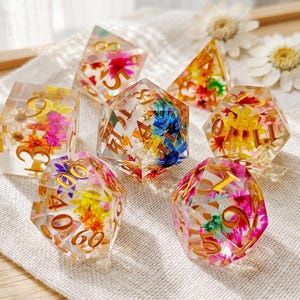 May include: A set of six polyhedral dice made of clear resin, each containing embedded colorful dried flowers and featuring gold-colored numbers. The collection includes various geometric shapes, such as a cube, a pyramid, and an icosahedron, all displaying vibrant floral patterns.