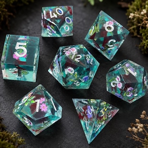 Pode incluir: Um conjunto de sete dados poliédricos feitos de resina transparente azul-petróleo, contendo flores em miniatura cor-de-rosa e roxas e folhagem verde. Os números estão gravados com um acabamento metálico prateado em cada face.