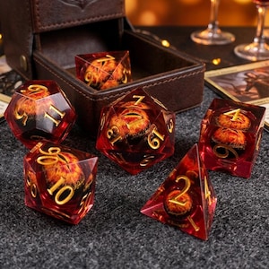 May include: A set of polyhedral dice with a translucent red resin finish and gold-colored numbers. Each die contains a suspended, fiery orange and yellow eye-like sphere. The set includes a d20, d12, d10, d8, d6, and d4, displayed near a brown box.