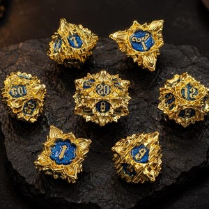 May include: A set of seven polyhedral dice featuring a metallic gold finish with intricate, sharp-edged floral or crystalline detailing. Each face contains a recessed blue enamel inlay with embossed gold numbers, including a twenty-sided die.