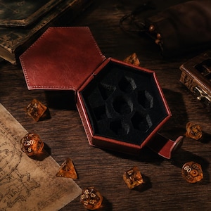 May include: A hexagonal dark red leather dice box with a black felt-lined interior featuring molded compartments for seven polyhedral dice. Several translucent amber-colored dice with gold numbering are scattered around the open container.