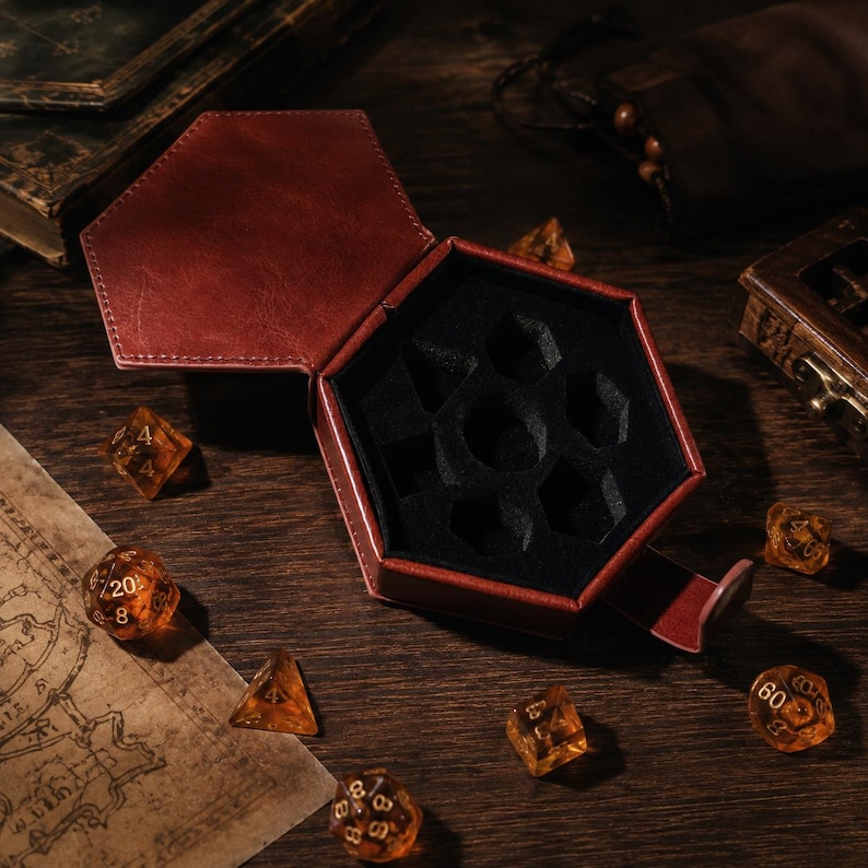 May include: A hexagonal dark red leather dice box with a black felt-lined interior featuring molded compartments for seven polyhedral dice. Several translucent amber-colored dice with gold numbering are scattered around the open container.