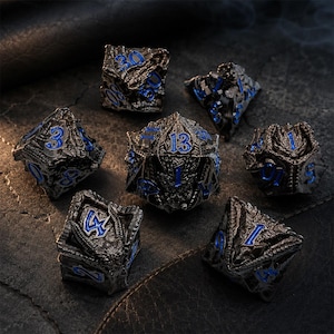 Seven-piece black metal DND dice set with vivid blue numbers and detailed dragon-inspired carvings. The polyhedral dice are arranged on a dark stone and leather surface with the D20 centered.