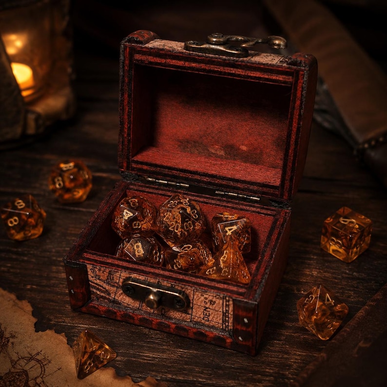 May include: A small, rectangular wooden chest with a dark red finish and a metal latch, containing several translucent amber-colored polyhedral dice with gold-colored numbers. Additional matching dice are scattered around the open chest.