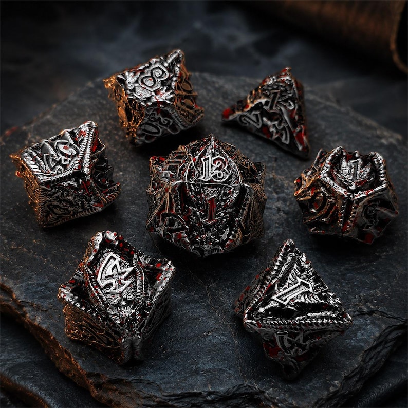 Seven-piece silver, black, and red dragon-themed metal DND dice set with raised numbers, sculpted scales, wings, and fantasy details. The polyhedral dice are arranged on a rugged dark stone slab.