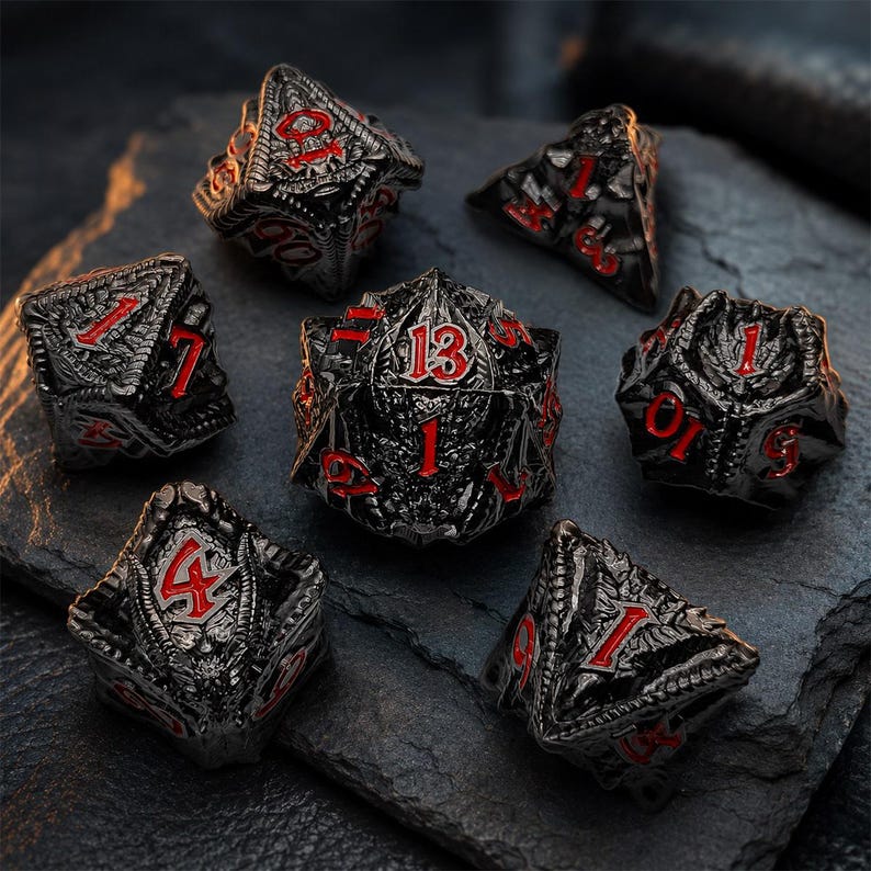 Seven-piece glossy black dragon-themed metal DND dice set with bold red numbers and sculpted scales, wings, and dragon details. The polyhedral dice are displayed on a dark textured stone surface.