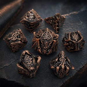 Seven-piece antique copper and black dragon-themed metal DND dice set featuring raised numbers, sculpted wings, scales, and fantasy details. The polyhedral dice are arranged on a rugged dark stone slab.