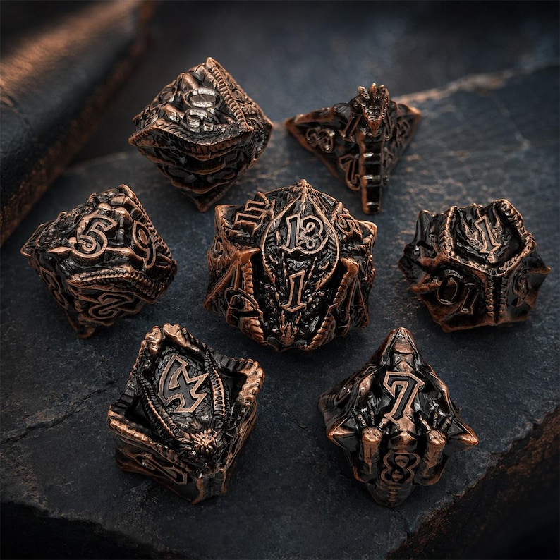 Seven-piece antique copper and black dragon-themed metal DND dice set featuring raised numbers, sculpted wings, scales, and fantasy details. The polyhedral dice are arranged on a rugged dark stone slab.