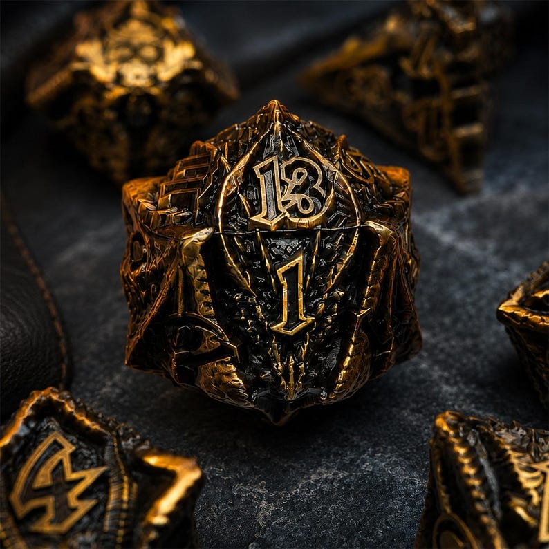 Close-up of a black and antique gold dragon-themed D20 featuring raised numbers 13 and 1, sculpted wings, scales, and dragon details. Matching metal dice appear softly blurred on a dark stone background.
