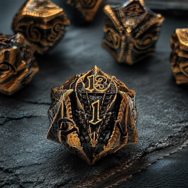 May include: A twenty-sided die featuring a dark black base with metallic gold highlights. The surface is sculpted with intricate dragon-like scales and wing textures. The numbers are embossed in a bold, stylized font, with the number one prominently displayed on the front face.