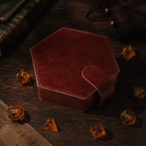 May include: A hexagonal dark red storage box with a textured surface and a matching strap closure. The box is surrounded by several translucent amber-colored polyhedral dice with gold-colored numbers, including a twenty-sided die.