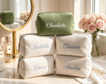 May include: A collection of five rectangular cosmetic bags with a waffle-textured fabric. One bag is sage green with the name Charlotte embroidered in white, while four cream-colored bags feature the names Isabella, Eleanor, Grace, and Amelia in blue.