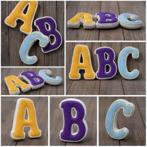 Op de afbeelding: Een set driedimensionale gehaakte letters A, B en C. De letter A is geel, de letter B is paars en de letter C is lichtblauw. Elke letter is voorzien van een witte gehaakte rand en een dikke, gevulde constructie voor een pluche uiterlijk.
