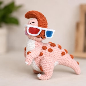 May include: A crochet dinosaur toy in light pink with a white belly and dark orange spots along its back and head. The dinosaur features a prominent orange crest and wears white-framed 3D glasses with one red lens and one blue lens on its face.
