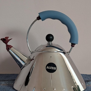 May include: A polished stainless steel kettle with a conical body, featuring a blue curved handle, a black spherical lid knob, and a dark red bird-shaped whistle on the spout. A circular black label with the white text ALESSI is on the front.