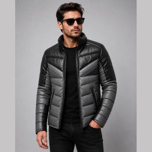 Könnte beinhalten: Eine farblich abgesetzte Steppjacke mit hohem Kragen, langen Ärmeln und einem Frontreißverschluss. Das Design umfasst schwarze und graue horizontal gesteppte Paneele mit einem kleinen dreieckigen Logo-Aufnäher im oberen Brustbereich.