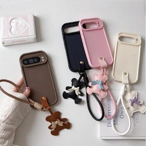 May include: Four rectangular smartphone cases in brown, black, pink, and cream, each featuring a matching wrist strap and a dangling teddy bear charm with a small bow. The cases have cutouts for camera lenses and are arranged on a flat surface.