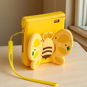 Könnte beinhalten: Eine leuchtend gelbe Schutzhülle für ein faltbares Smartphone mit einem erhabenen, dreidimensionalen Bienenmotiv auf der Rückseite. Die Biene hat gelb-braune Streifen und hellgelbe Flügel. Ein passendes gelbes Handgelenkband ist befestigt.