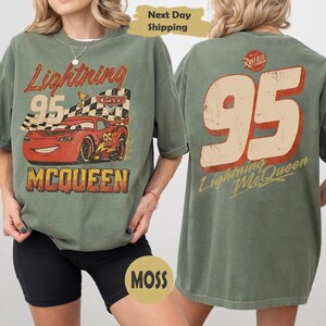 May include: An olive green t-shirt featuring a graphic of a red race car with the number 95 and a checkered flag on the front. The back of the shirt displays the large number 95 in a distressed font with the text Lightning McQueen below it.