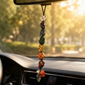 May include: A hanging car ornament featuring a vertical string of seven raw, multi-colored gemstones encased in black netting. The stones are arranged in a gradient from white to purple, blue, green, yellow, orange, and red, ending in a cluster of small charms.