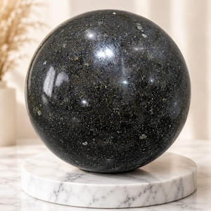 Puede incluir: Una esfera de piedra pulida con una superficie oscura y moteada que presenta motas de color verde oscuro, negro y tonos dorados metálicos. La esfera tiene un acabado liso y reflectante que capta la luz, creando brillos intensos en su superficie.