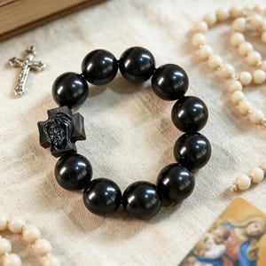 May include: A circular bracelet composed of large, smooth, polished black beads. One bead is shaped as a square cross featuring a detailed, raised relief carving of a face. The bracelet is arranged in a round formation on a textured, light-coloured fabric surface.