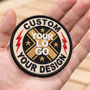 Puede incluir: Un parche bordado circular con un diseño de cruz en negro y dorado con cuatro pequeñas estrellas doradas. El borde es crema con texto negro que dice CUSTOM YOUR LOGO YOUR DESIGN y dos rayos rojos a los lados. El texto central es blanco.