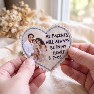 Può includere: Una toppa in tessuto beige a forma di cuore con bordi dentellati, che presenta una fotografia stampata di un uomo, una donna e un bambino. Il testo ricamato in blu scuro recita: MY PARENTS WILL ALWAYS BE IN MY HEART 5-7-05, con cuciture blu.