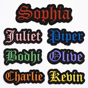 Pode incluir: Sete emblemas bordados com nomes, bordas pretas e letras góticas. Os nomes incluem Sophia em vermelho, Juliet em rosa, Piper em azul, Bodhi em verde, Olive em roxo, Charlie em laranja e Kevin em amarelo, todos sobre tecido preto.