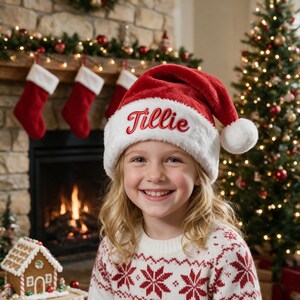 Op de afbeelding: Een rood-witte pluche kerstmuts met een witte rand van imitatiebont waarop de naam Tillie in rode schrijfletters is geborduurd. De muts heeft een witte pompon aan de punt en is gemaakt van zachte, pluizige stof in een klassieke kegelvorm.