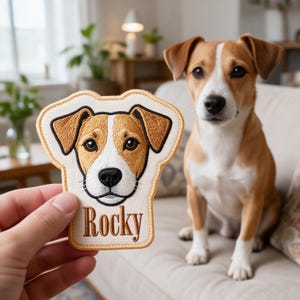 May include: An embroidered fabric patch featuring a brown and white Jack Russell Terrier dog face with the name Rocky stitched in brown thread below the dog's head. The patch has a light cream background and a tan border.