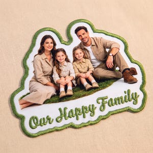 Könnte beinhalten: Ein individuell bestickter Aufnäher mit einem gedruckten Foto einer vierköpfigen Familie in beigefarbener Kleidung, die auf grünem Gras sitzt. Der Aufnäher hat einen dicken, grünen Stickrand und den Text Our Happy Family in grünem Garn unten.