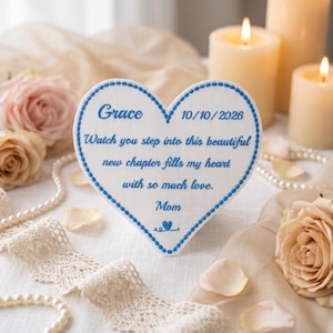 May include: A white heart-shaped plaque with a blue stitched border and blue embroidered text that reads: Grace 10/10/2026 Watch you step into this beautiful new chapter fills my heart with so much love. Mom, with a small blue heart icon.