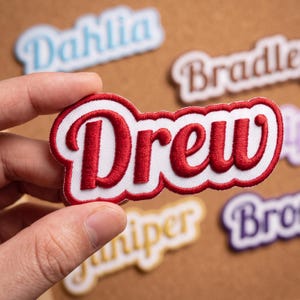 May include: An embroidered patch featuring the name Drew in bold red script lettering with a white background and a red border. The patch has a scalloped edge design and is held up against a background of other similar name patches.