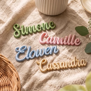 Könnte beinhalten: Vier personalisierte Namensaufnäher in Schreibschrift, jeder mit einer erhabenen, gepolsterten Textur. Die Namen Simone in Grün, Camille in Pink, Elowen in Hellblau und Cassandra in Gold sind in einer Anordnung auf strukturiertem Stoff.