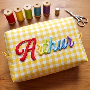 Puede incluir: Un estuche de tela con estampado de cuadros vichy en amarillo y blanco que presenta el nombre Arthur bordado en letras cursivas multicolores. El estuche está rodeado por cinco carretes de hilo de madera en rojo, amarillo, verde, azul y morado.
