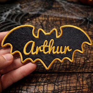 Op de afbeelding: Een zwarte geborduurde patch in de vorm van een vleermuis met een gouden rand en de naam Arthur in gouden cursieve letters in het midden. De patch heeft een textuurrijk stoffen oppervlak en een duidelijk vleermuissilhouet met vleugels.