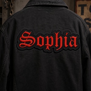 May include: A black jacket featuring a large, embroidered patch on the back. The patch displays the name Sophia in bold, red, gothic-style lettering with a black outline, set against the dark fabric of the garment's back panel and collar.