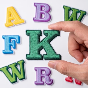 May include: A collection of embroidered alphabet letter patches in various colors including yellow, purple, green, blue, and red. Each letter features a bold, block-style font with a white border. One dark green letter K is held in the foreground.