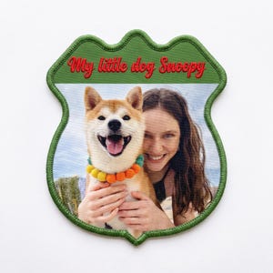 Peut inclure: Un écusson brodé en forme de bouclier avec une bordure verte présentant une photographie imprimée d'une personne souriante tenant un chien beige et blanc portant un collier à pompons colorés. Le texte My little dog Snoopy est brodé en rouge.