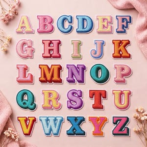 Può includere: Una collezione di ventisei toppe ricamate con le lettere dell'alfabeto, disposte su cinque righe. Ogni lettera presenta un carattere in stile blocco con un effetto tridimensionale, realizzato in vari colori vivaci tra cui rosa, blu, giallo, arancione e viola.