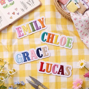 Peut inclure: Quatre écussons brodés portant les prénoms EMILY, CHLOE, GRACE et LUCAS dans des couleurs variées. Chaque lettre est cousue individuellement avec une bordure blanche, créant un style d'étiquette nominative distinct pour chaque étiquette en tissu.