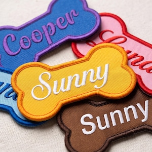 Peut inclure: Une collection d'écussons brodés en forme d'os de différentes couleurs, notamment bleu, jaune, rose et marron. Chaque écusson présente un texte personnalisé cousu avec un fil contrastant, affichant des noms comme Cooper et Sunny.
