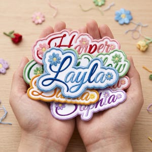 May include: A collection of embroidered name patches featuring scalloped edges and small floral accents. Each patch displays a unique name in cursive script, including Layla in blue, Harper in red, and Sophia in purple, with various thread colours.