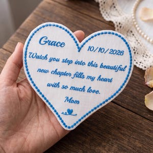 Può includere: Una toppa in tessuto bianco a forma di cuore con bordi ricamati in blu e testo blu che recita: Grace, 10/10/2026, Watch you step into this beautiful new chapter fills my heart with so much love, Mom, e una piccola icona a forma di cuore.