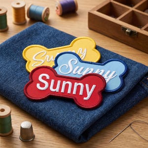 Peut inclure: Trois écussons en tissu en forme d'os de couleur jaune, bleu clair et rouge, chacun portant le mot Sunny brodé en fil blanc. Les écussons sont superposés sur un morceau de tissu denim bleu foncé plié, avec des fournitures de couture.
