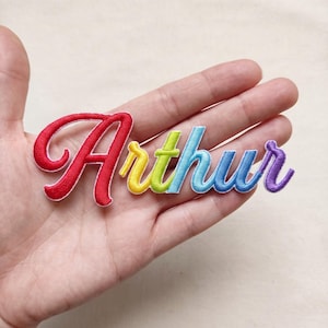 Könnte beinhalten: Ein gestickter Aufbügler mit dem Namen Arthur in einer Schreibschrift. Die Buchstaben zeigen einen Regenbogen-Farbverlauf, beginnend mit Rot für das große A, gefolgt von Gelb, Grün, Hellblau und Violett.